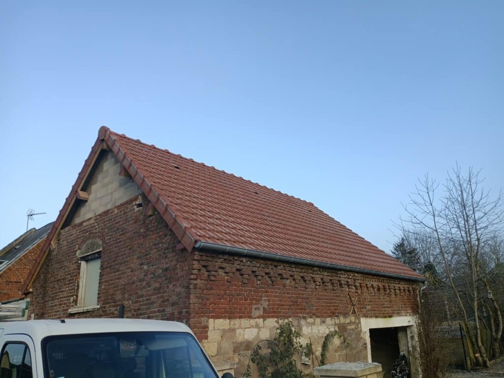 Toit de tuiles rouges rénové sur une vieille grange en briques avec gouttière et partie supérieure en parpaings, ciel bleu clair.